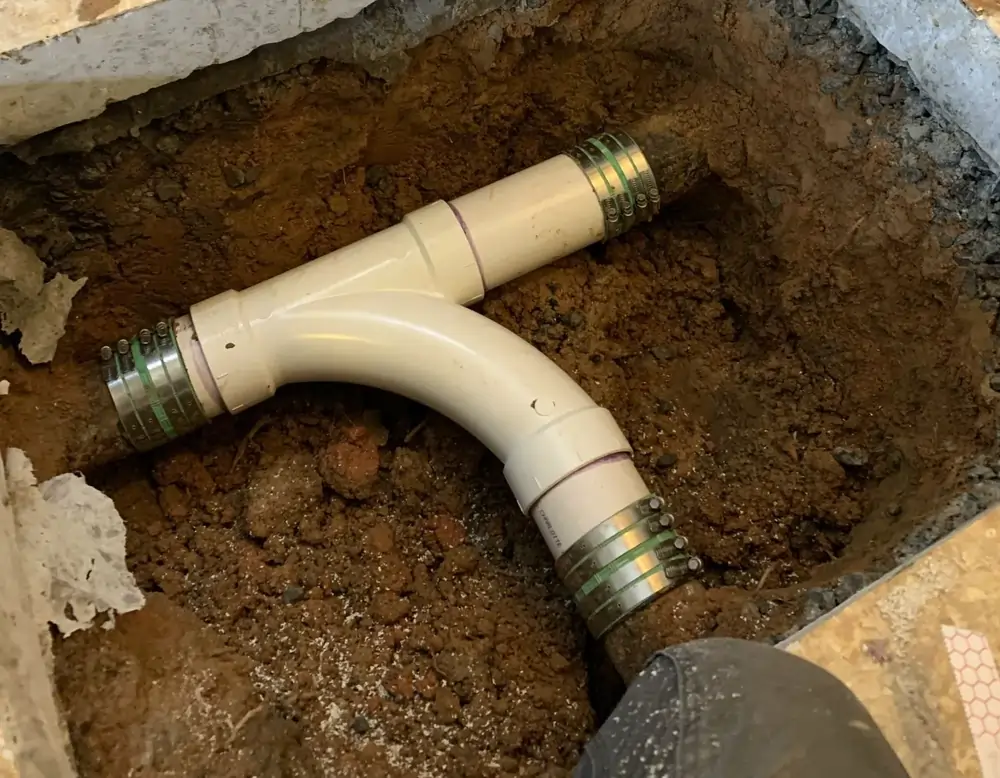 A pipe junction with metal clamps is visible in an excavated dirt area, showing plumbing installation work in progress.