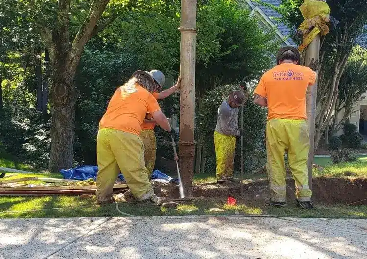 Four workers in orange shirts and yellow pants dig around a large pipe in a lush, green outdoor setting.