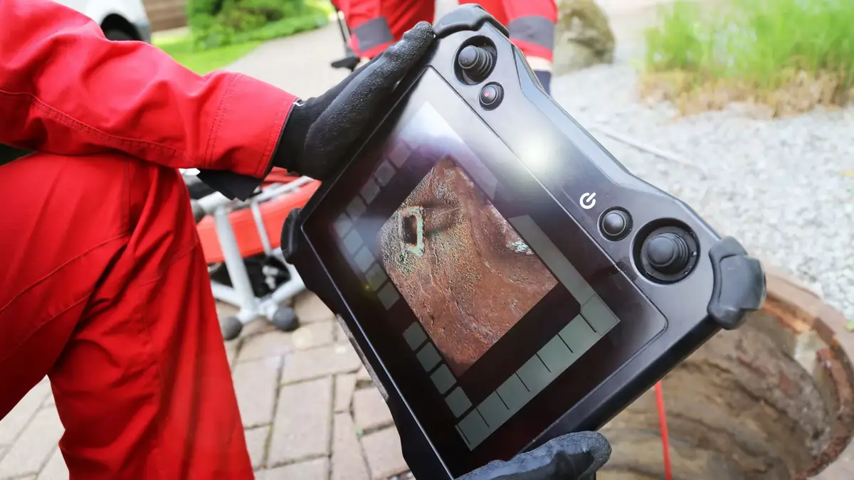 A person in red overalls, wearing black gloves, holds a screen displaying an underground pipe inspection image.