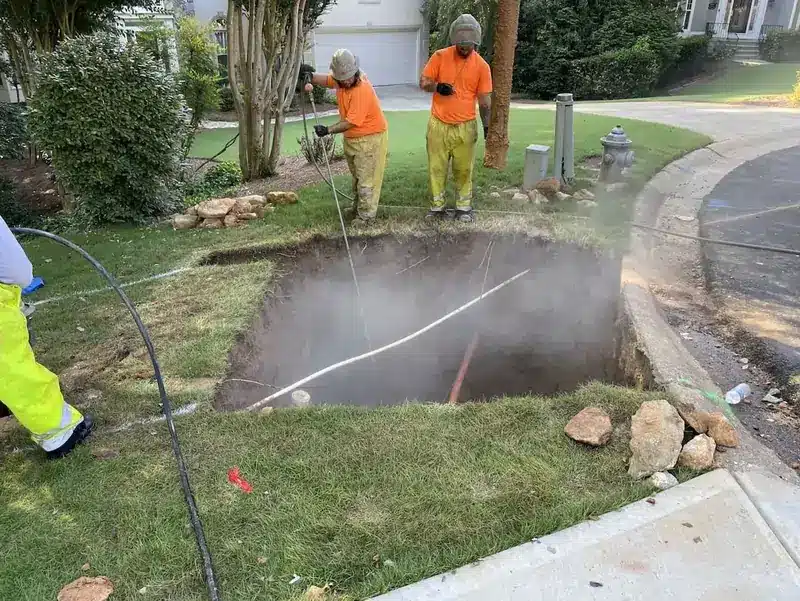Two workers in orange shirts and helmets inspect a large, steaming sinkhole on a grassy roadside.