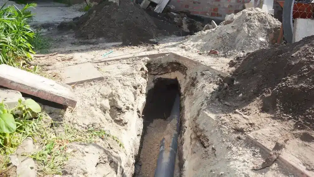 Deep trench in a garden under construction with a visible black pipe buried in the ground, surrounded by soil piles and greenery on the side.