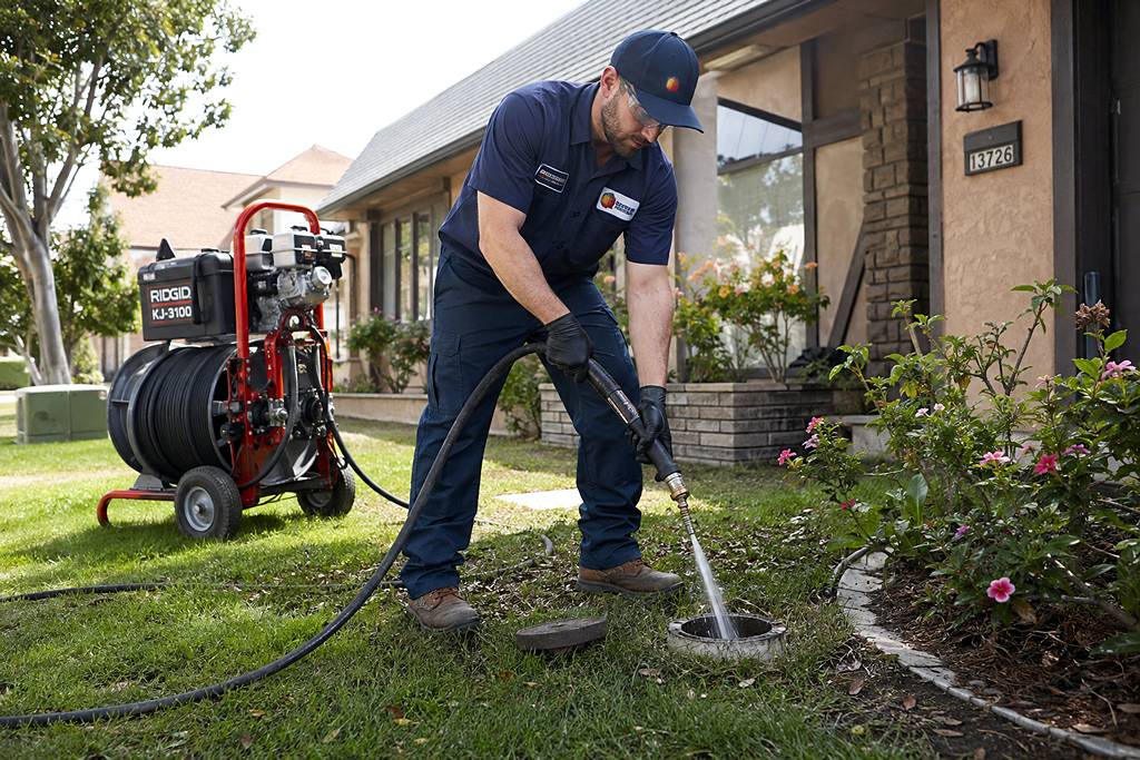 A plumber operating a hydro jetting machine connected to a residential sewer clean-out access point in a yard.